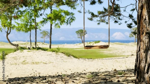 Rope swing hangs on tree on a beautiful sunny beach with white sand and blue water of lake Baikal. Swing shake on a pine tree against the background of the mountain landscape in summer day