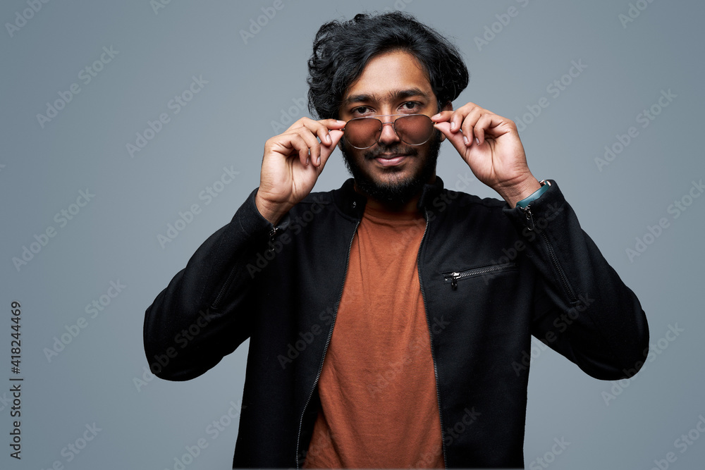Obraz premium Wearing black jacket and orange shirt hipster person takes off his sunglasses looking at camera and posing in gray background.