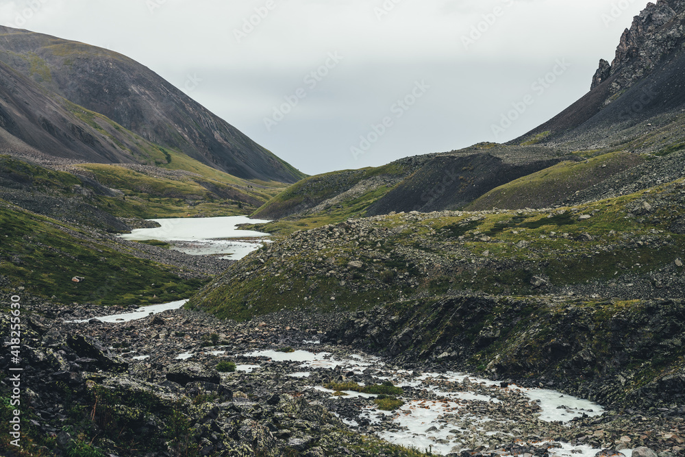 Atmospheric landscape with mountain lake and mountain creek among moraines in rainy weather. Bleak overcast scenery with milky river and lake among rocks. Gloomy view to milk mountain river and lake. Atmospheric landscape with mountain lake and mountain creek among moraines in rainy weather. Bleak overcast scenery with milky river and lake among rocks. Gloomy view to milk mountain river and lake.