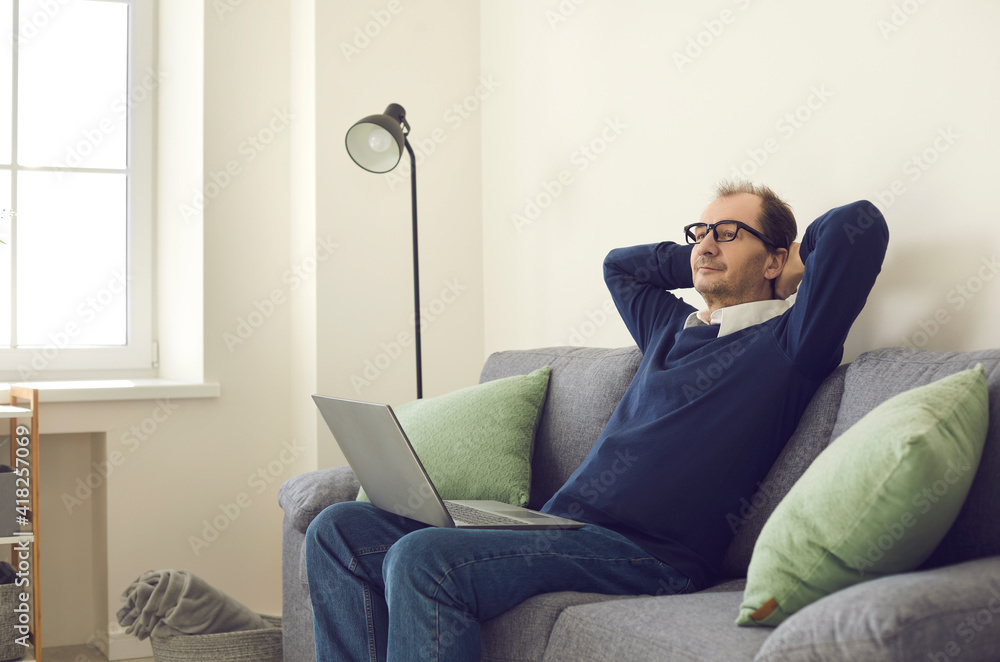Relaxed middle-aged businessman took a break from work sitting on a comfortable sofa with a laptop and putting his hands behind his head. Concept is no stress, fatigue and relaxation.