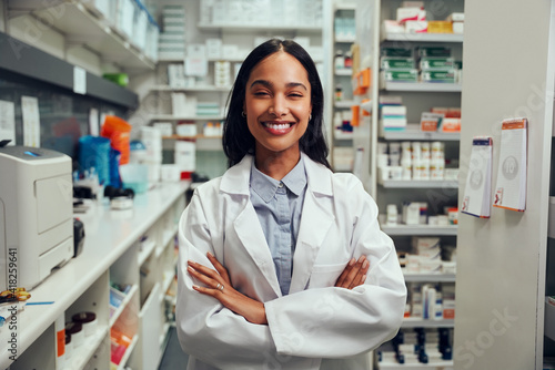 Cheerful young african american female pharmacist wearing labcoat with folded hands standing in chemist near shelves photo