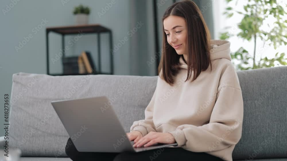 Pretty woman in casual hoodie sitting on comfy couch with portable laptop on knees and typing on keyboard. Female freelancer doing remote work from home. Pretty woman in casual hoodie sitting on comfy couch with portable laptop on knees and typing on keyboard. Female freelancer doing remote work from home.