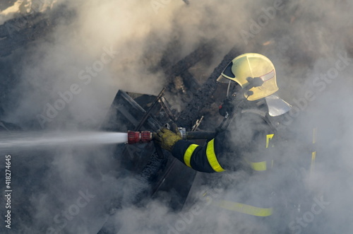 Firefighter with a breathing apparatus shrouded and surrounded by thick smoke extinguish the fire of wooden buildings with water from a hose