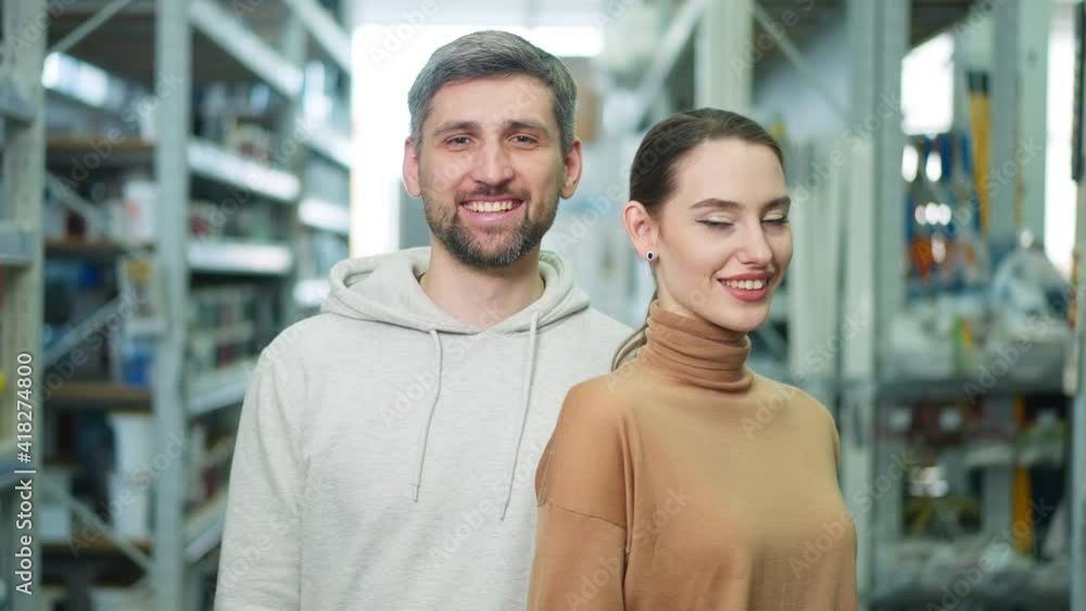 Portrait of happy young satisfied couple posing in hardware store indoors. Beautiful woman and handsome man looking at camera smiling standing between rows of good in shop. Portrait of happy young satisfied couple posing in hardware store indoors. Beautiful woman and handsome man looking at camera smiling standing between rows of good in shop.