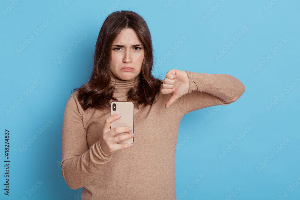 Fototapeta premium Displeased young brown haired woman casual beige turtleneck holds mobile phone, showing thumb down isolated on blue color background, frowning face and pout lips.