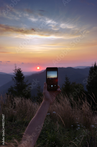 Arm of young man with a mobile phone on landscape background. Tourist taking picture of beautiful sunrise in mountain. Hands with smart phone. A man takes a photo for memory at dawn.