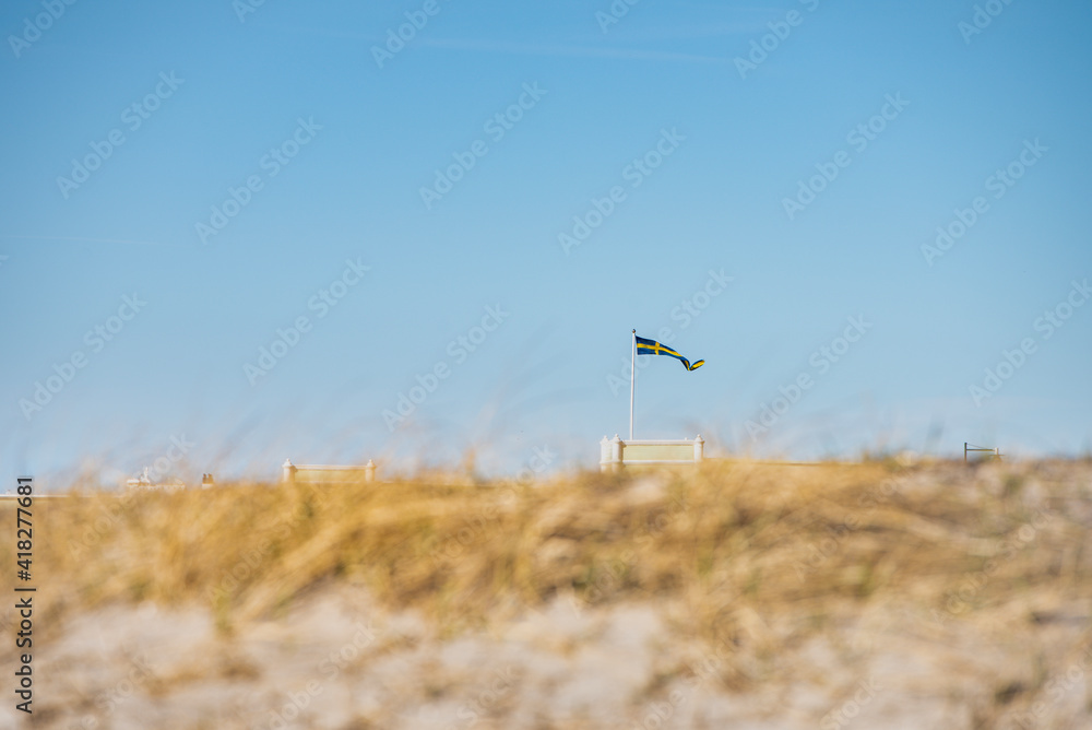 Swedish flag fluttering on a pole, captured through the sandy dunes of Ribersborgsstranden (Ribersborg Strand or Beach). Scandinavian Swedish banderole waving on Ribersborgs Kallbadhus - Malmo, Sweden Swedish flag fluttering on a pole, captured through the sandy dunes of Ribersborgsstranden (Ribersborg Strand or Beach). Scandinavian Swedish banderole waving on Ribersborgs Kallbadhus - Malmo, Sweden