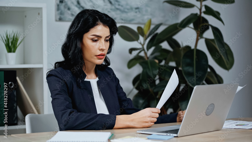 Businesswoman looking at paper near laptop on blurred foreground in office Businesswoman looking at paper near laptop on blurred foreground in office