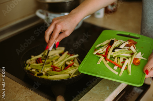 Fresh vegetables cut into strips: cherry tomato, zucchini, red pepper, eggplant. Girl's hand close up throws them from the board to the pan on the stove to fry and cook a delicious meal