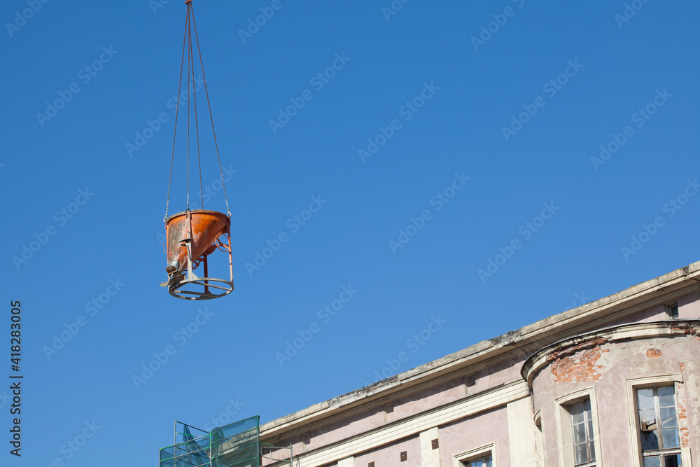 Hanging orange concrete mixer against the blue sky and a fragment of a renovated building.
