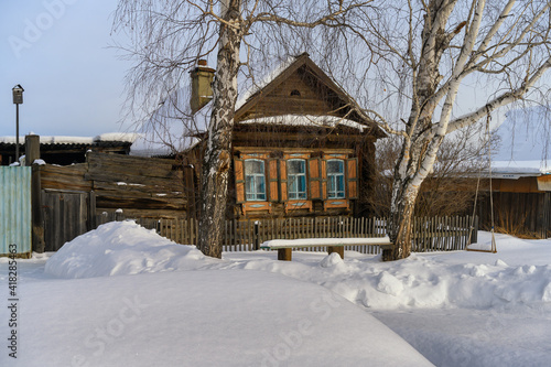 One-storey wooden old house in the village in the traditional Russian style in winter. High birches grow near the house, there is a low board fence, a bench, a swing. Pure fluffy snow on a sunny winte
