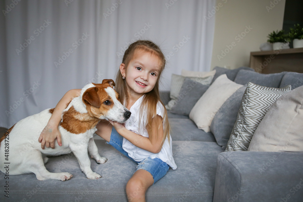 Happy preschooler girl hugs her dog Jack Russell Terrier breed at home on the couch. Quarantine. Best friends are resting and having fun at home in the room. Animal games Happy preschooler girl hugs her dog Jack Russell Terrier breed at home on the couch. Quarantine. Best friends are resting and having fun at home in the room. Animal games