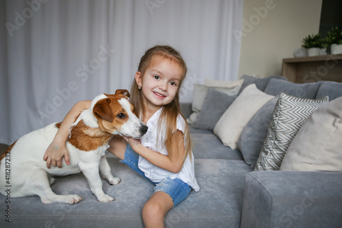 Happy preschooler girl hugs her dog Jack Russell Terrier breed at home on the couch. Quarantine. Best friends are resting and having fun at home in the room. Animal games