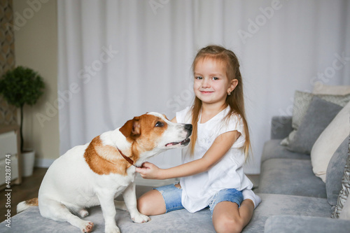 Happy preschooler girl hugs her dog Jack Russell Terrier breed at home on the couch. Quarantine. Best friends are resting and having fun at home in the room. Animal games © Valeriya