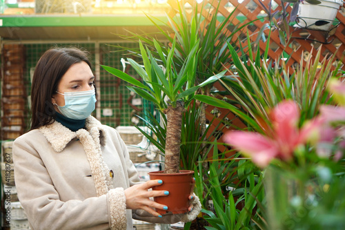 Young woman wearing in a face medical mask buying a jucca palm in a garden center. Coronavirus time. photo