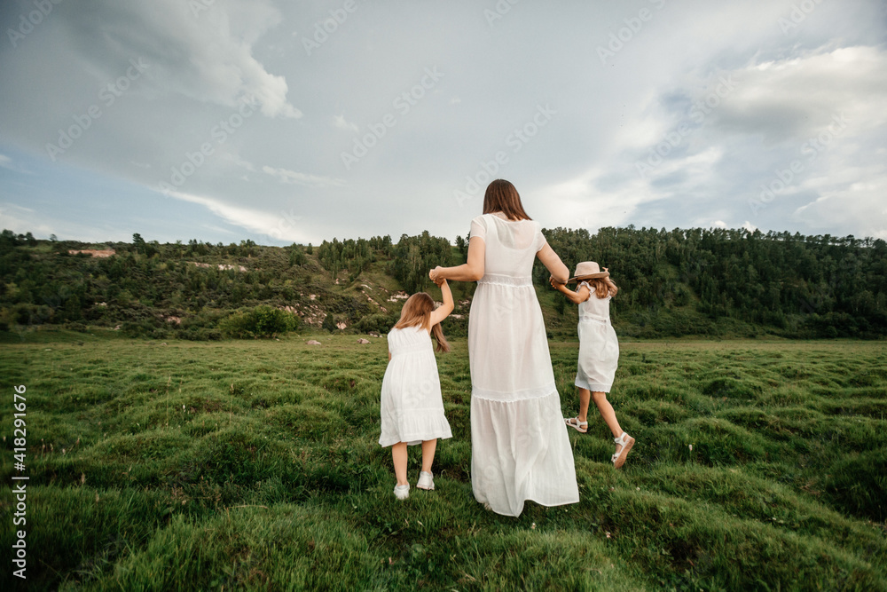 Happy young family: mother and three daughters in white clothes spending time together outside in green nature. Rear view