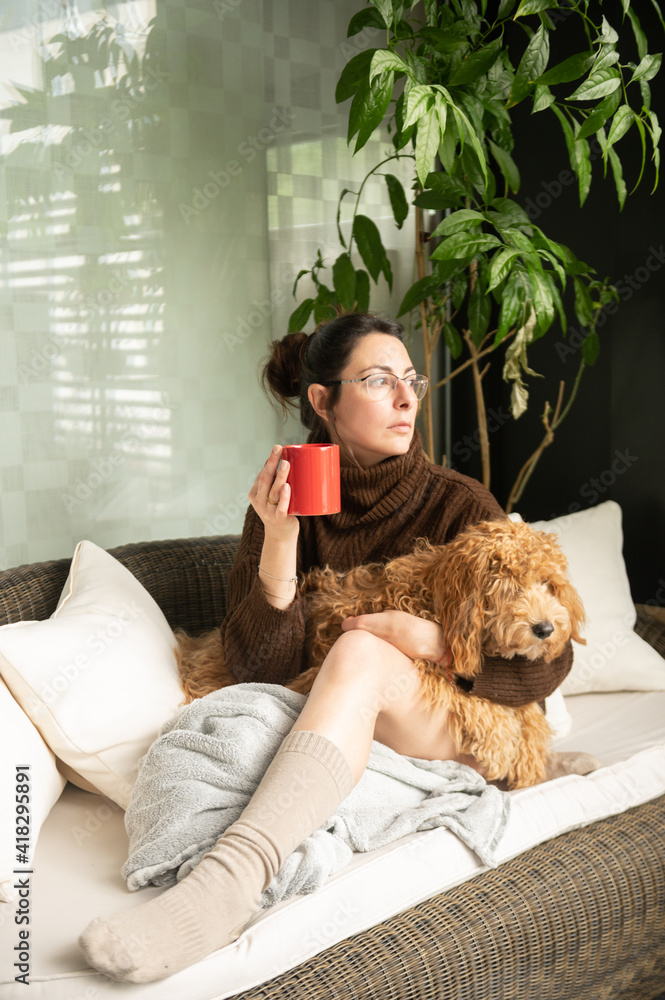 Young and attractive girl sitting comfortably on a couch hugging her adorable dog while holding a cup of coffee and contemplating life through the window of her house. Young and attractive girl sitting comfortably on a couch hugging her adorable dog while holding a cup of coffee and contemplating life through the window of her house.