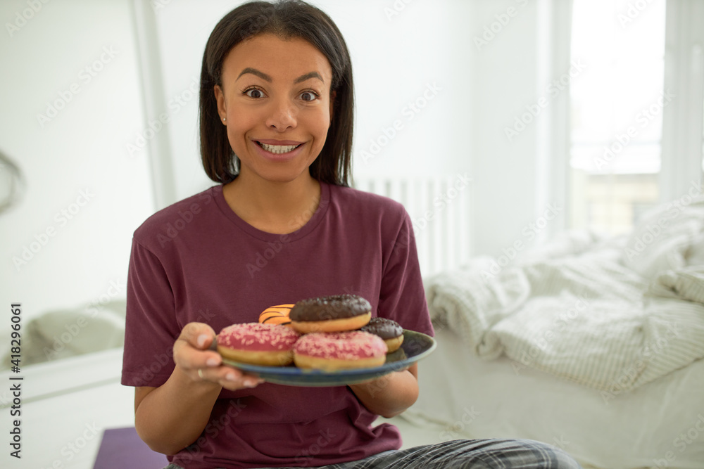 Portrait of excited overjoyed young dark skinned woman with slim body enjoying sweet sugary dessert in the morning, holding plate of doughnuts with colorful glazing, looking at camera in excitement Portrait of excited overjoyed young dark skinned woman with slim body enjoying sweet sugary dessert in the morning, holding plate of doughnuts with colorful glazing, looking at camera in excitement