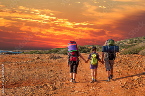 Dad, daughter and mom are holding hands at sunset along a dirt road towards the Mediterranean Sea on the island of Cyprus