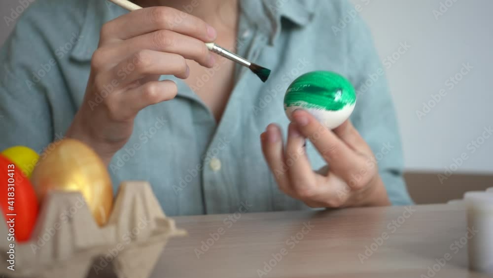 Young woman paints egg with green paint next to cardboard egg stand with different colorful eggs. Prepare for Easter, happy Easter concept. Young woman paints egg with green paint next to cardboard egg stand with different colorful eggs. Prepare for Easter, happy Easter concept.