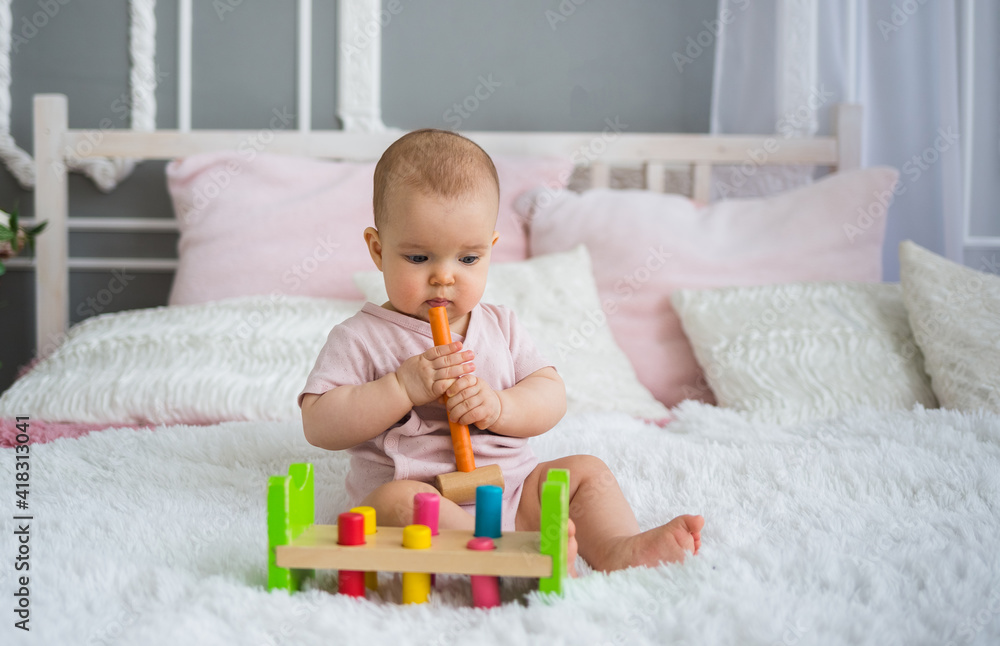 Naklejka premium a beautiful baby girl in a pink bodysuit sits on the bed and plays with an educational toy