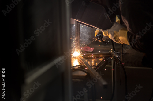 a welder in a welding mask, gloves welds metal with a welding machine and an electrode in his hand, sparks fly, a beautiful glow, bokeh, orange light on a black background, a flash of light and smoke