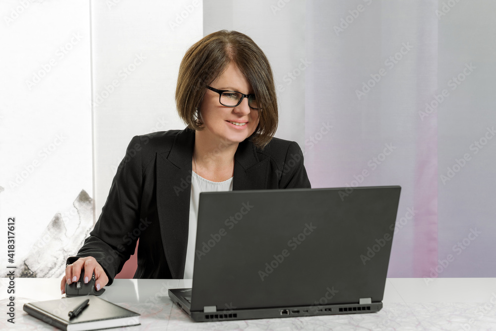 A brunette bussiness woman in glasses in a black jacket works on the computer in the office from home with smile A brunette bussiness woman in glasses in a black jacket works on the computer in the office from home with smile