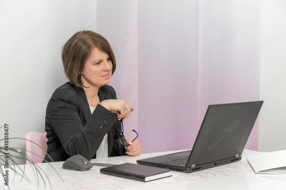 A brunette bussiness woman in a black jacket works on the computer in the office and holding glasses in his hands with smile A brunette bussiness woman in a black jacket works on the computer in the office and holding glasses in his hands with smile
