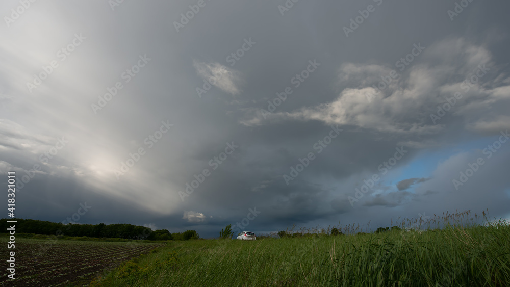 Rain clouds are a strip of deciduous forest and green grass at the edge of a field and a white car, panoramic landscape.