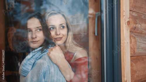 Young lesbian couple on winter vacation. Hugging and looking through the window on snow covered neighborhood. High quality photo © CameraCraft