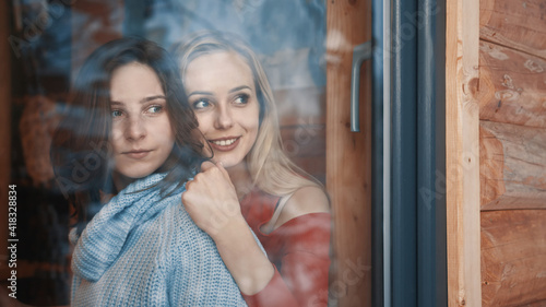 Young lesbian couple on winter vacation. Hugging and looking through the window on snow covered neighborhood. High quality photo