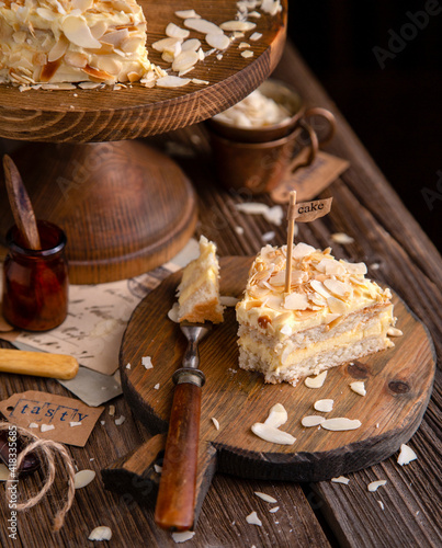 tasty homemade slice of cake with two biscuit layers, white cream and almond flakes on wooden cake stand on rustic table