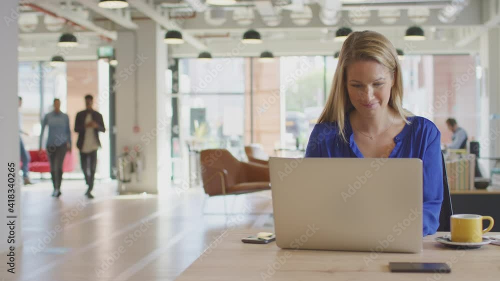 Dolly shot of casually dressed young businesswoman working on laptop at desk in modern open plan start up office with colleagues in background - shot in slow motion Dolly shot of casually dressed young businesswoman working on laptop at desk in modern open plan start up office with colleagues in background - shot in slow motion