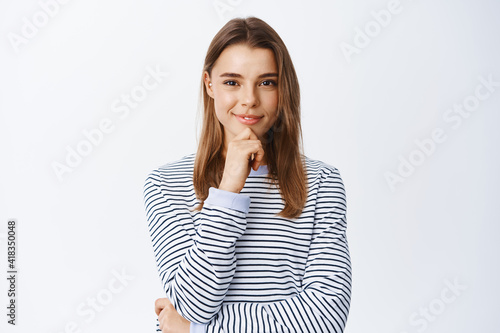 Hmm interesting. Thoughtful blond woman having an idea, listening and looking at camera confident, smiling pleased of good plan, standing pensive over white background © Cookie Studio