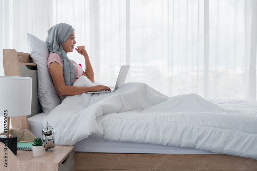Young Asian woman wearing a hijab uses smartphone and laptop at her desk. Attaching a pink ribbon represents recovery from a breast cancer patient. Breast cancer concept, cancer prevention concept. Young Asian woman wearing a hijab uses smartphone and laptop at her desk. Attaching a pink ribbon represents recovery from a breast cancer patient. Breast cancer concept, cancer prevention concept.