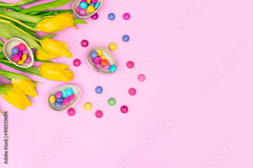 Easter border composition. Chocolate eggs, colorful candies bonbons, yellow tulips on pink background. Stylish decor minimal concept. Text space. Festive flat lay greeting card