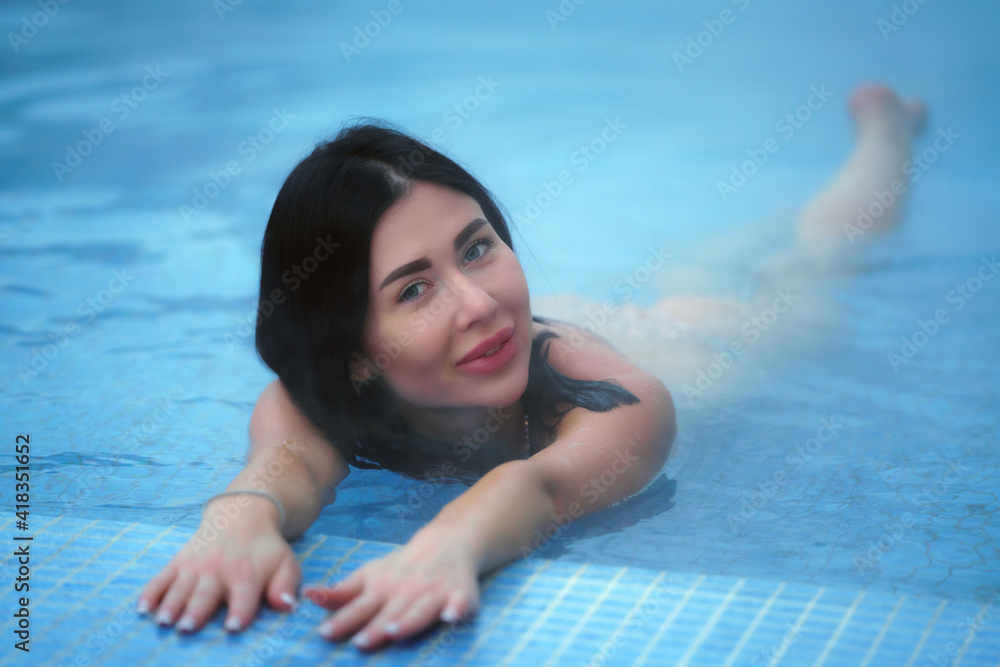 Adult brunette relaxation lies in geothermal water in pool at spa resort and looking at camera. Soft selective focus on model's eyes.
