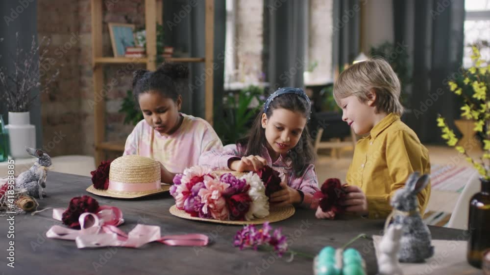 Medium shot of cute children sitting together at table and decorating straw hats with flowers for Easter Medium shot of cute children sitting together at table and decorating straw hats with flowers for Easter