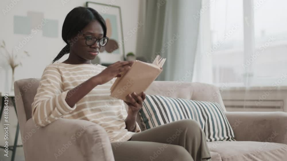 Medium shot of young African-American woman wearing casual clothes and glasses sitting on couch in living room and reading interesting book Medium shot of young African-American woman wearing casual clothes and glasses sitting on couch in living room and reading interesting book