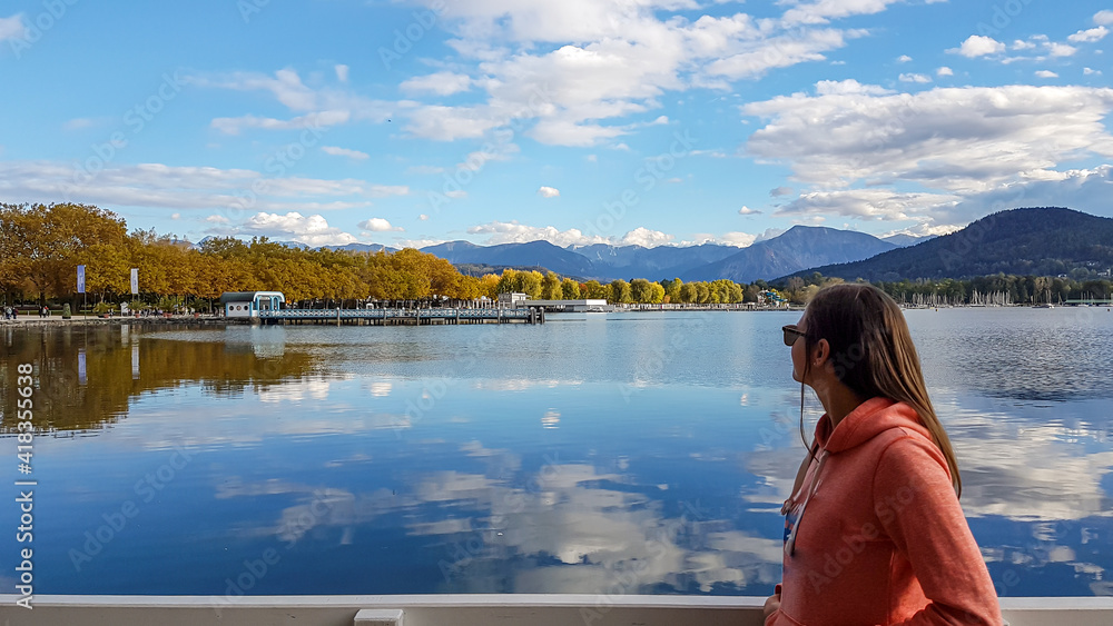 A woman in orange hoodie walking along a pier on the Woerthersee in Austria. The calm lake's surface is reflecting the surrounding mountains and clouds. Alpine landscape. The woman enjoys the peace.