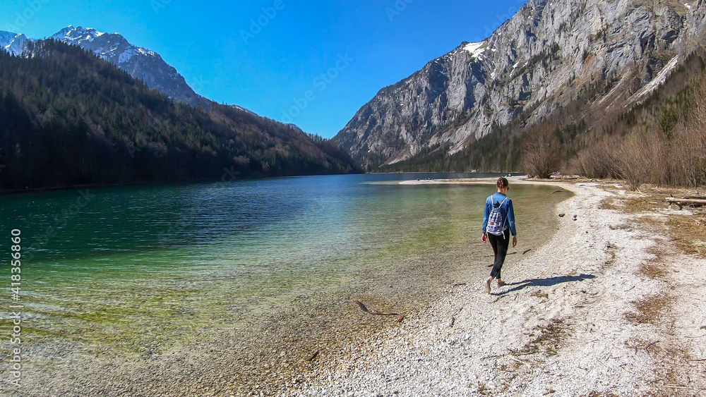 A woman walking along the shore of Leopoldsteiner lake in Austria. The lake is surrounded by high Alps. The shallow water is crystal clear, spring water has a calm surface. Early spring. Serenity