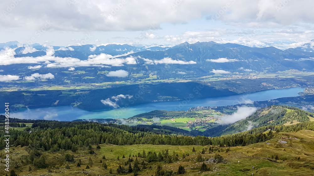 A panoramic view on the Millstaettersee from Granattor in Austrian Alps. The distant lake is surrounded by high mountains. There are few clouds above the lake and the valley. Endless mountain chains
