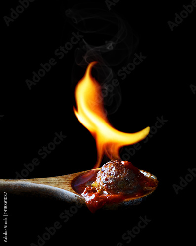 meatball with spicy tomato sauce in wooden spoon with fire shaped as pepper side view on black background isolated closeup. Selective focus