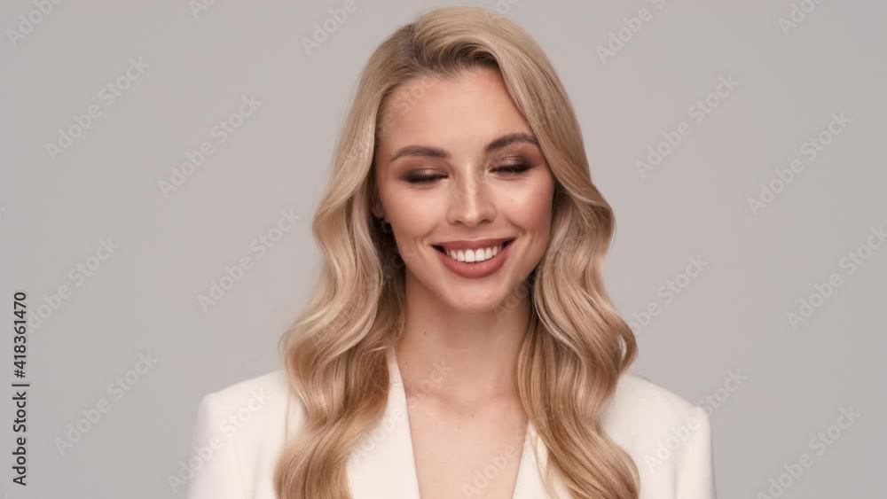 A dreaming young blonde businesswoman is smiling to the camera standing isolated over gray background in studio A dreaming young blonde businesswoman is smiling to the camera standing isolated over gray background in studio