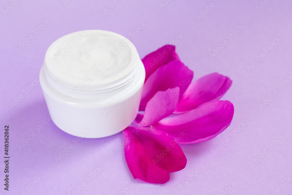 White moisturizing cream with peony extract and petals nearby it on pink background White moisturizing cream with peony extract and petals nearby it on pink background