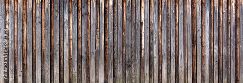 Panorama Holzwand aus vertikalen, grau verwitterten Brettern mit aufgesetzten braunen Rundhölzern