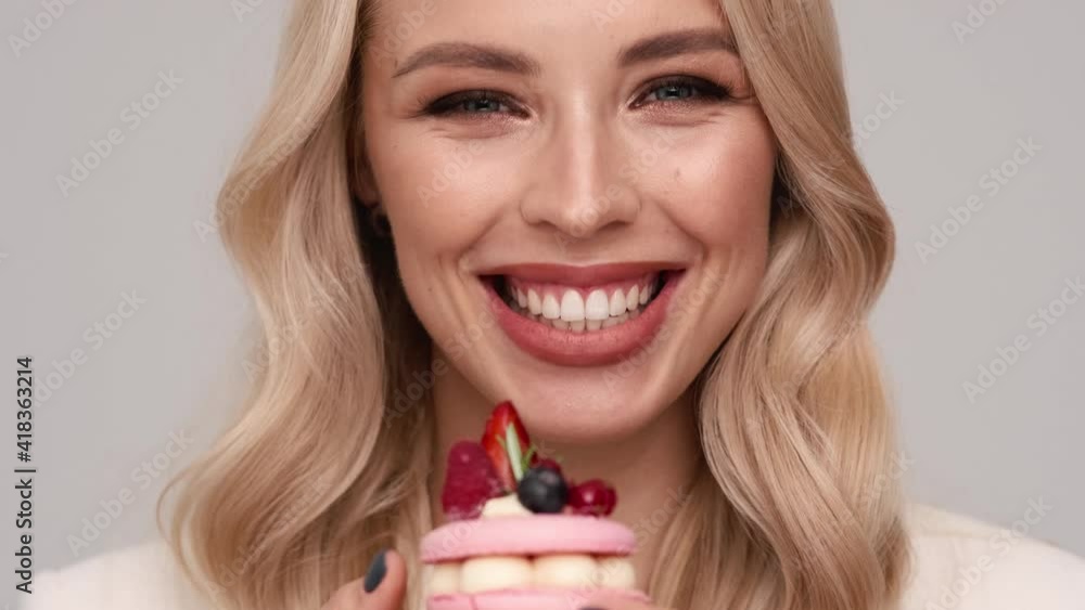 A close-up view of a smiling young blonde woman is presenting a cake standing isolated over gray background in studio A close-up view of a smiling young blonde woman is presenting a cake standing isolated over gray background in studio