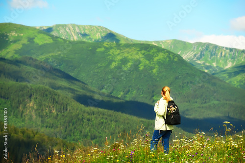 Girl tourist with a backpack for walks admires the views in the mountains. beautiful mountain summer green landscape with green mountains and blooming meadow.  © Ann Stryzhekin