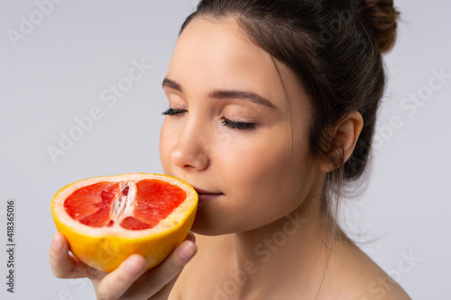 Portrait of a young girl with closed eyes enjoying the aroma of fresh, juicy grapefruit on a gray background. Enlarged photo.