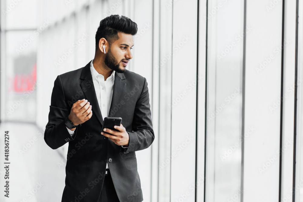 Handsome bearded Indian man, businessman, worker in black suit, he speaks on the phone through wireless headphones and looks out the window, office